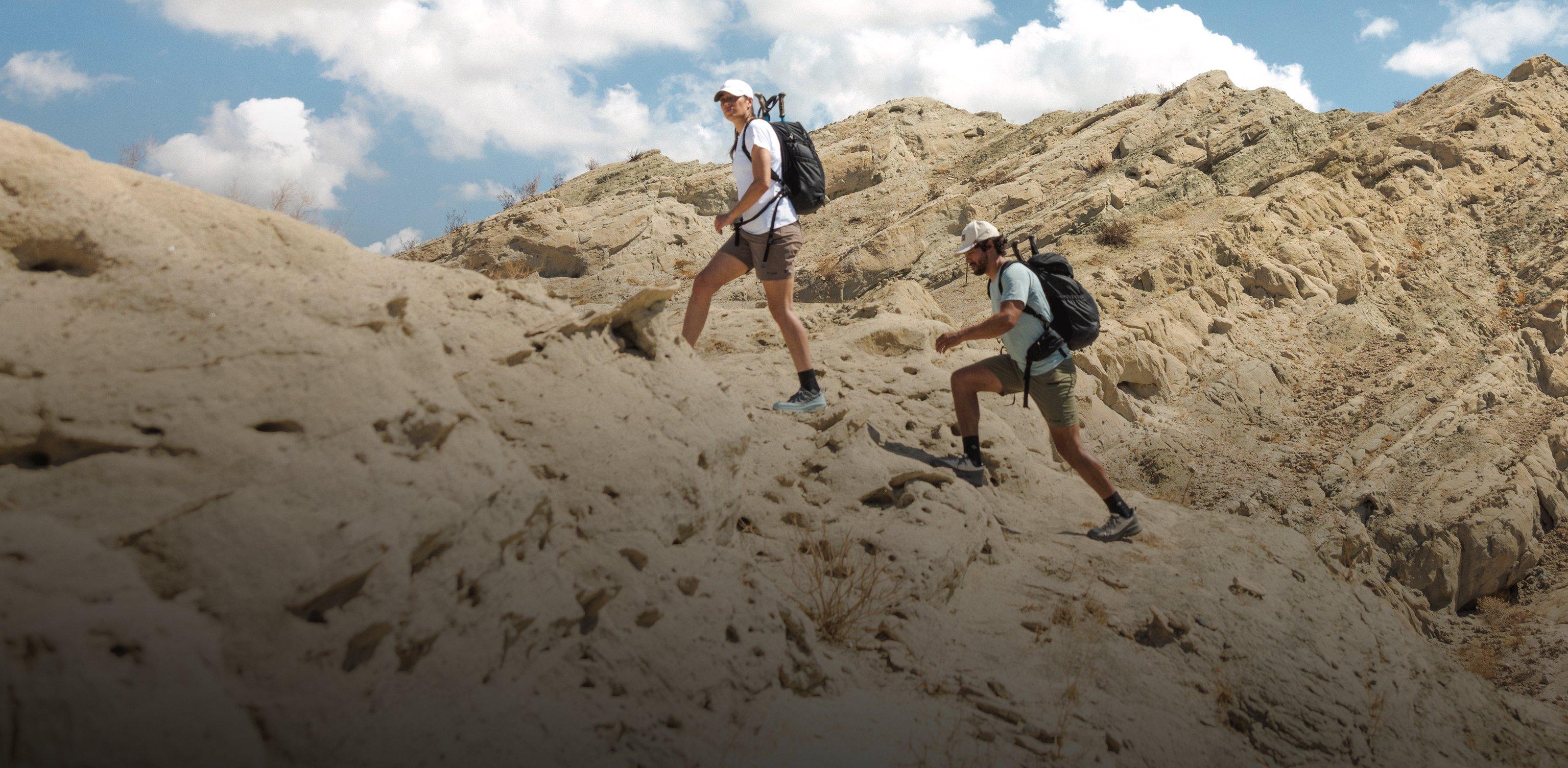 Two hikers with backpacks trekking across rocky desert terrain under a blue sky with clouds.