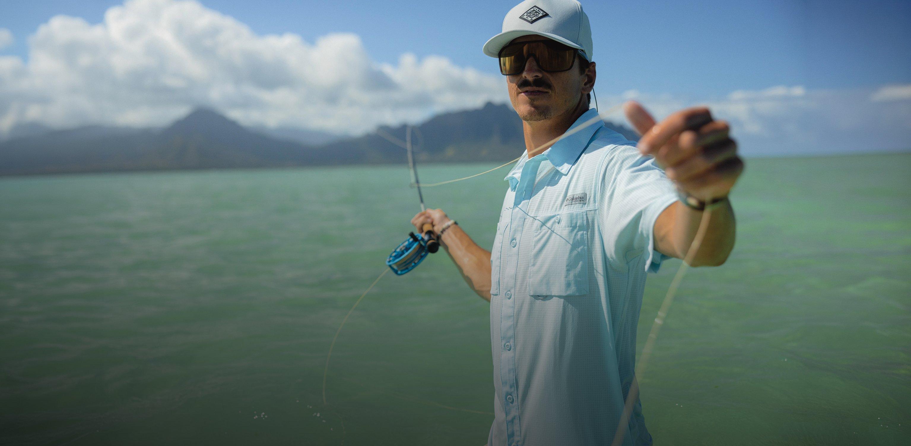 Man fly fishing in coastal waters wearing light blue shirt, cap, and sunglasses with mountains in background.