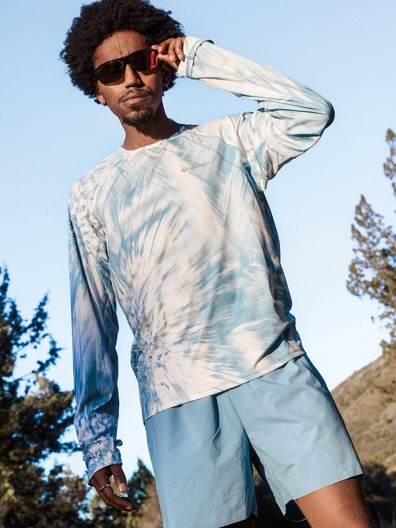 Man wearing sunglasses and blue tie-dye long-sleeve shirt outdoors, hand adjusting sunglasses.