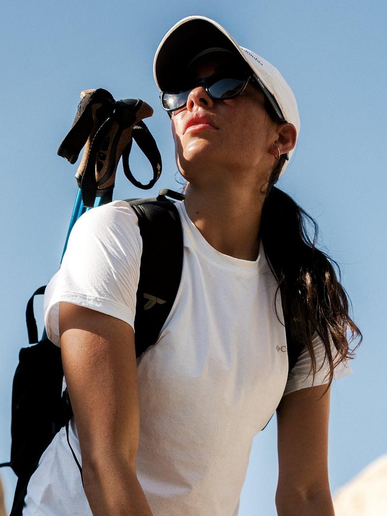 Woman wearing white t-shirt, cap, and sunglasses carries backpack and cycling gloves outdoors.