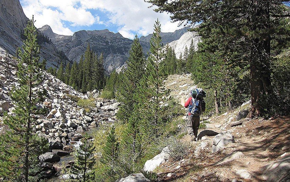A hiker with a large pack and trekking poles walks alongside a rocky creek toward large granite peaks.