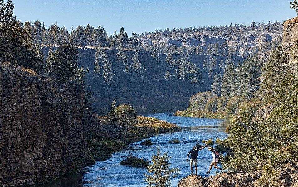 One person helps another jump from rock to rock along a river.