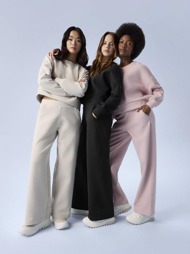 Three women standing together wearing matching sweatshirt and wide-leg pants sets in beige, black, and pink.