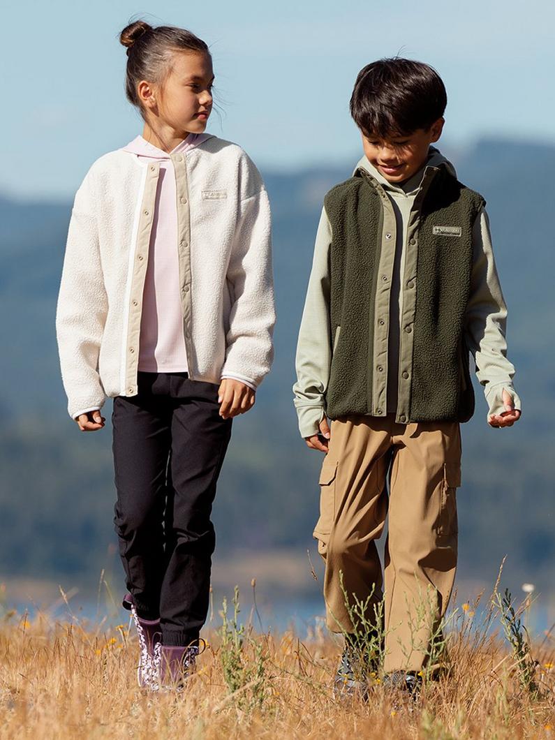 Two children walking through a grassy field wearing fleece vests and outdoor clothing.