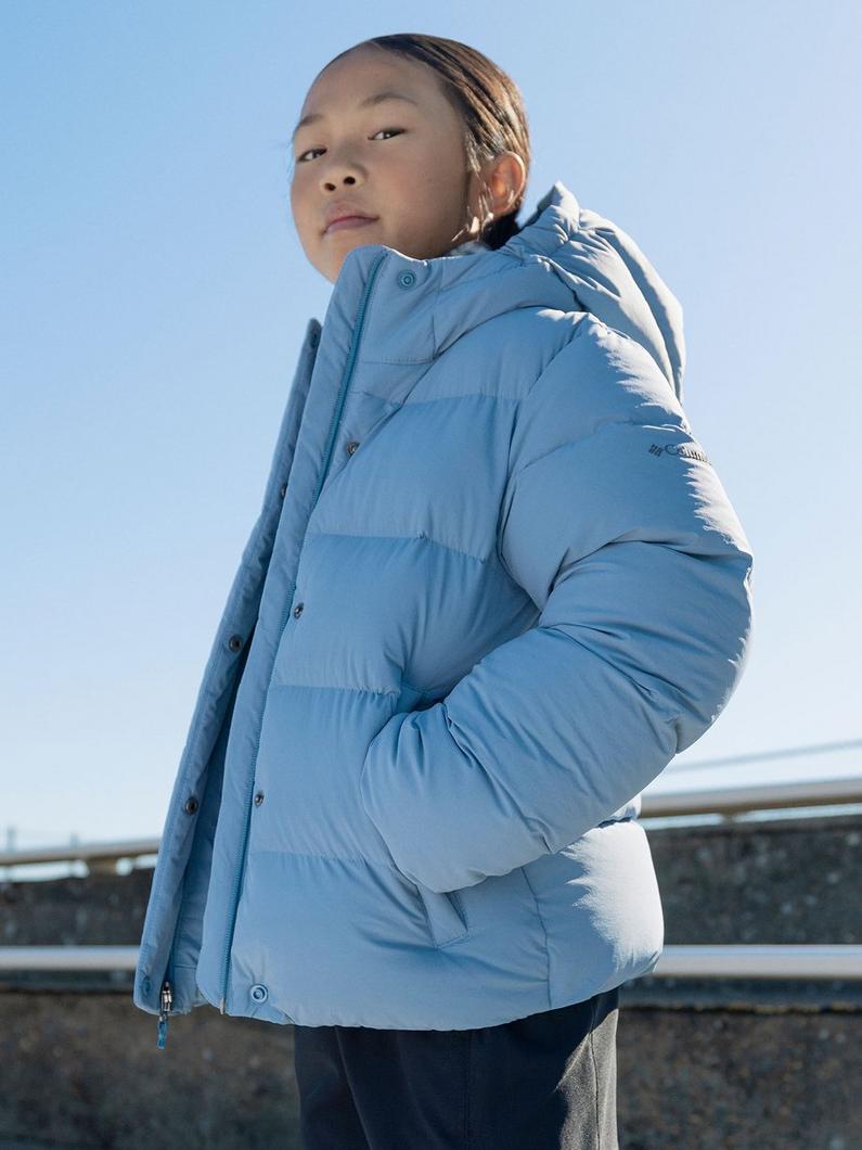 Child wearing blue quilted puffer jacket with hood, standing outdoors with hand in pocket.