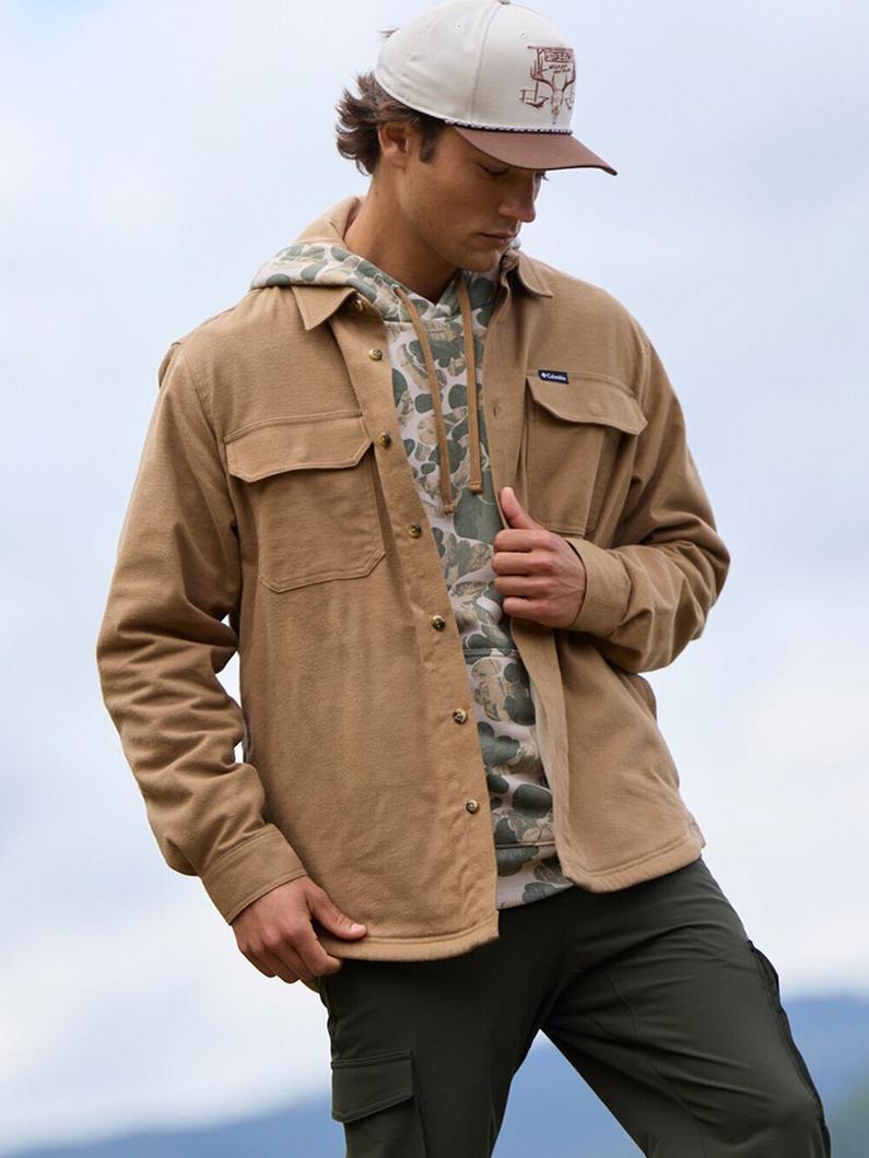 Man wearing tan fleece shirt jacket with chest pockets over camouflage hoodie and baseball cap.