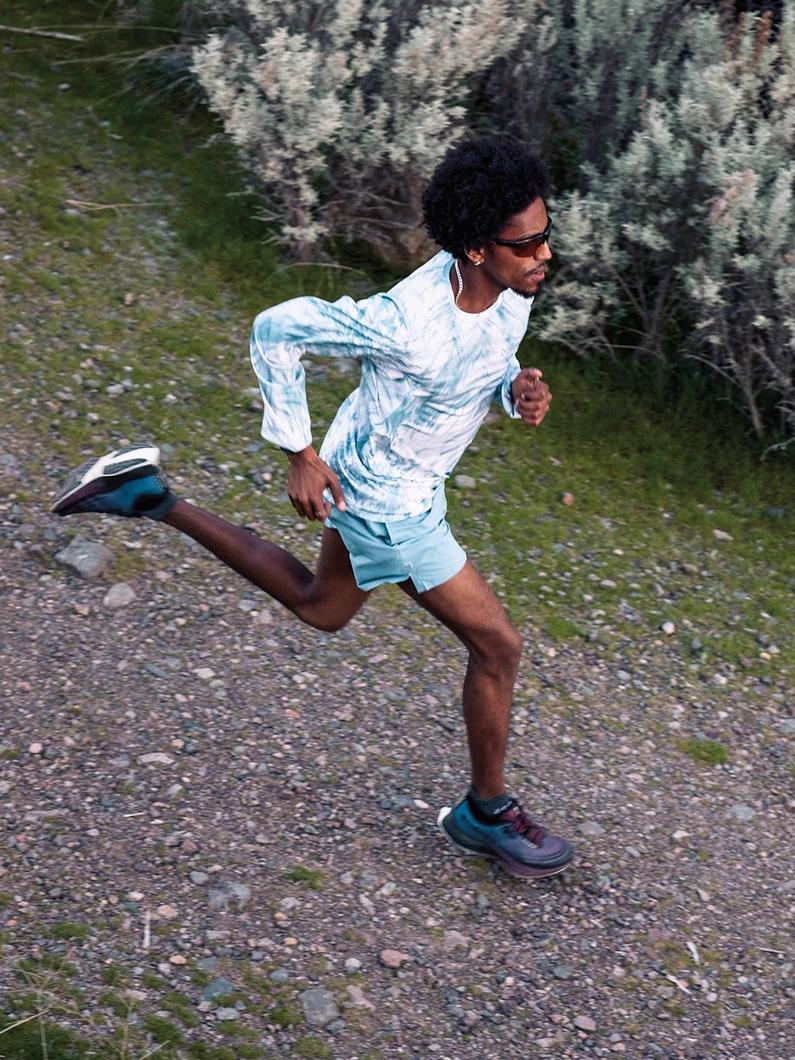 Man running on gravel trail wearing blue tie-dye long sleeve shirt, light blue shorts, and sneakers.