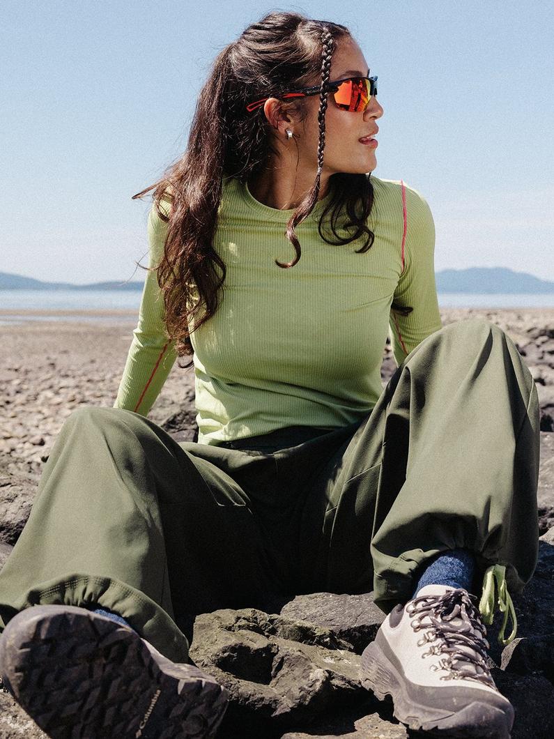 Woman sitting on beach rocks wearing lime ribbed long-sleeve top, olive pants, and orange-tinted sunglasses.