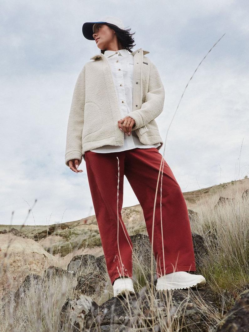 Woman standing on rocky terrain wearing cream sherpa jacket, white shirt, rust wide-leg pants, and white sneakers.