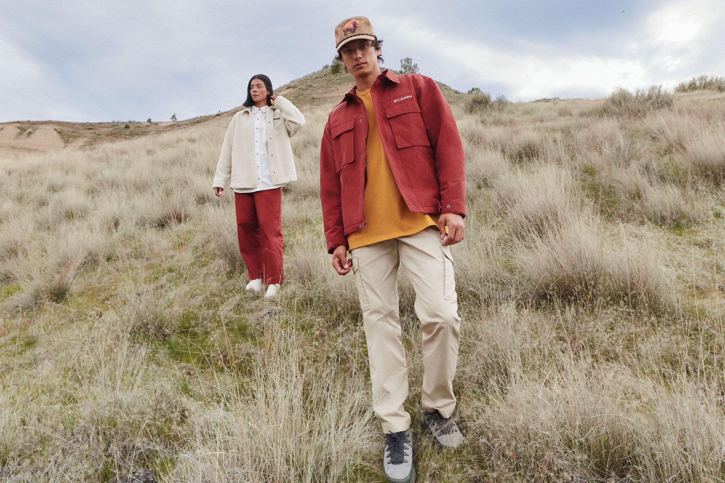 Man in red jacket and tan cap hiking through grassland with woman in cream jacket following behind.