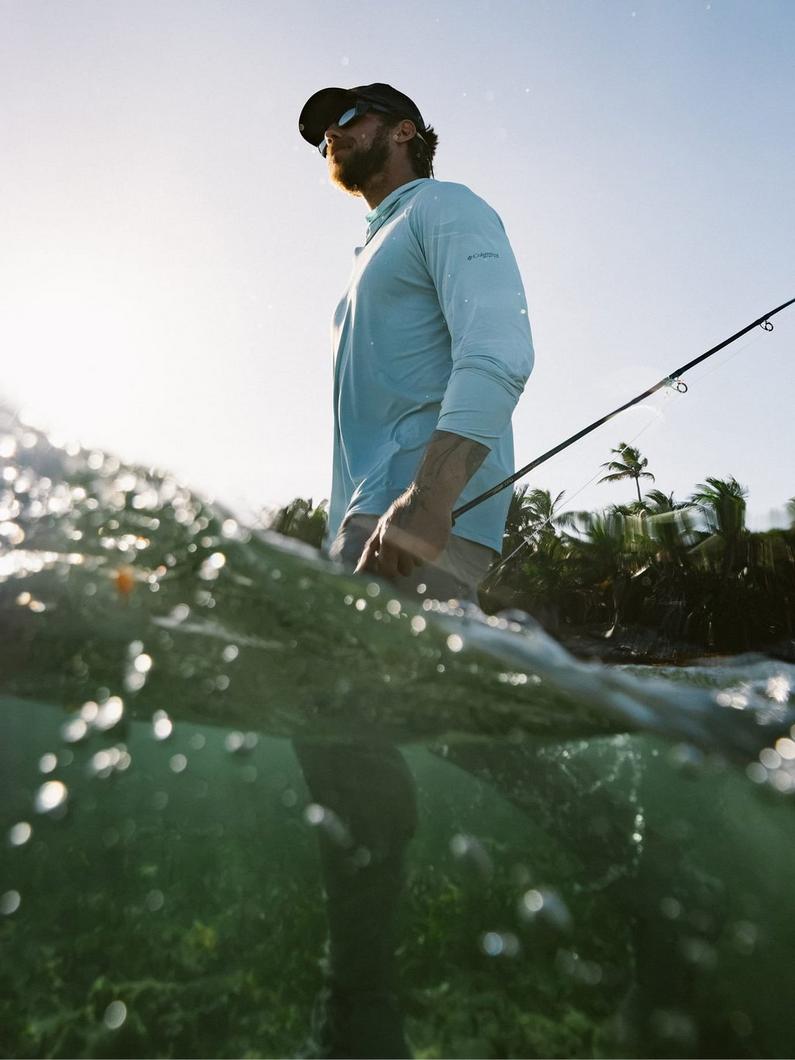 Man wearing light blue fishing shirt and cap holding fishing rod while standing in tropical waters.