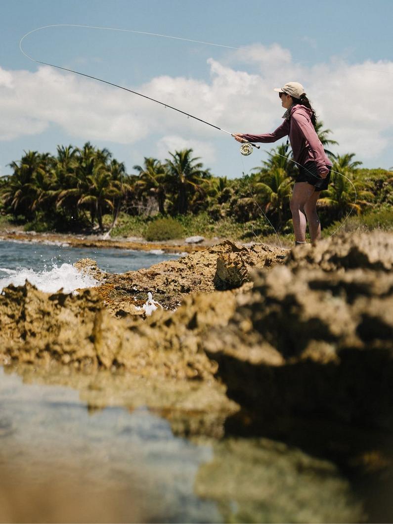 Woman fly fishing from rocky coastal outcrop with palm trees in tropical setting.