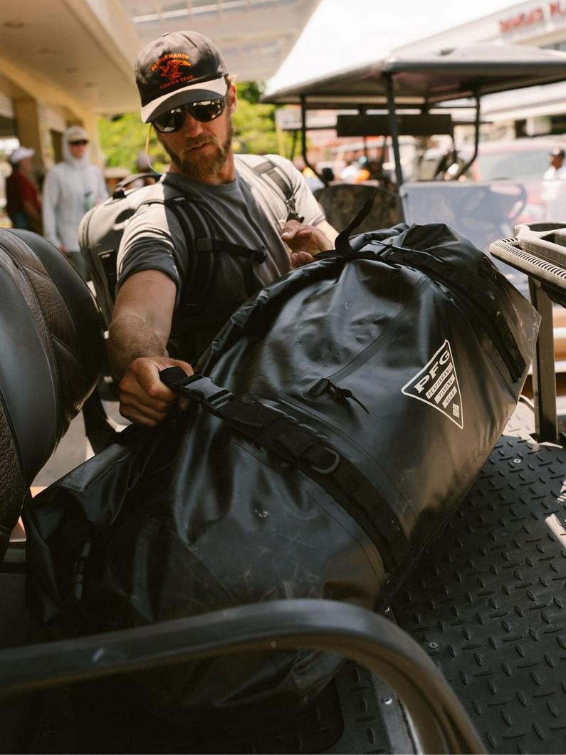 Man wearing cap and sunglasses loading a large black duffel bag onto a golf cart.