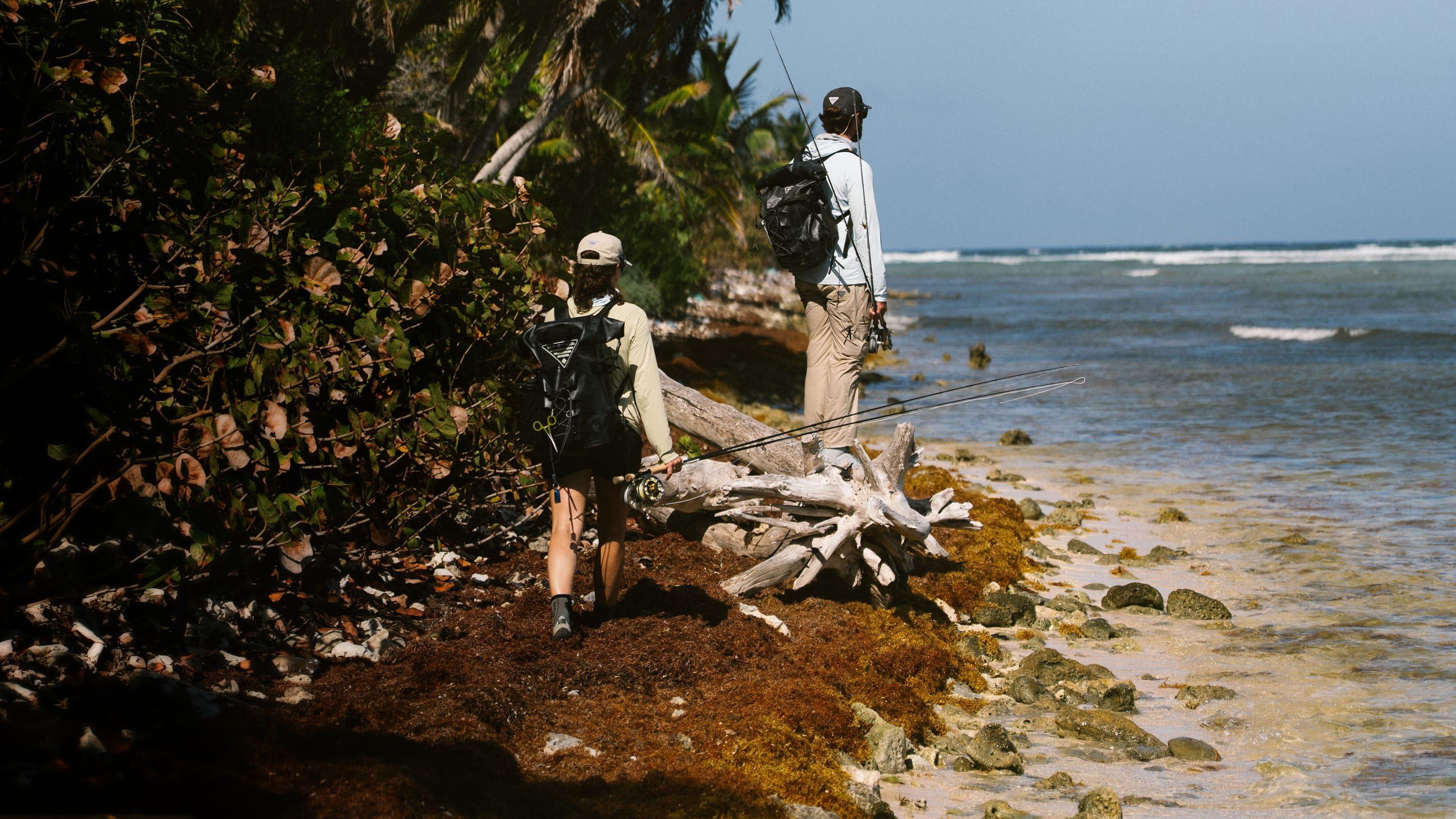 Two anglers fishing from a rocky tropical shoreline with backpacks and rods near driftwood and palm trees.