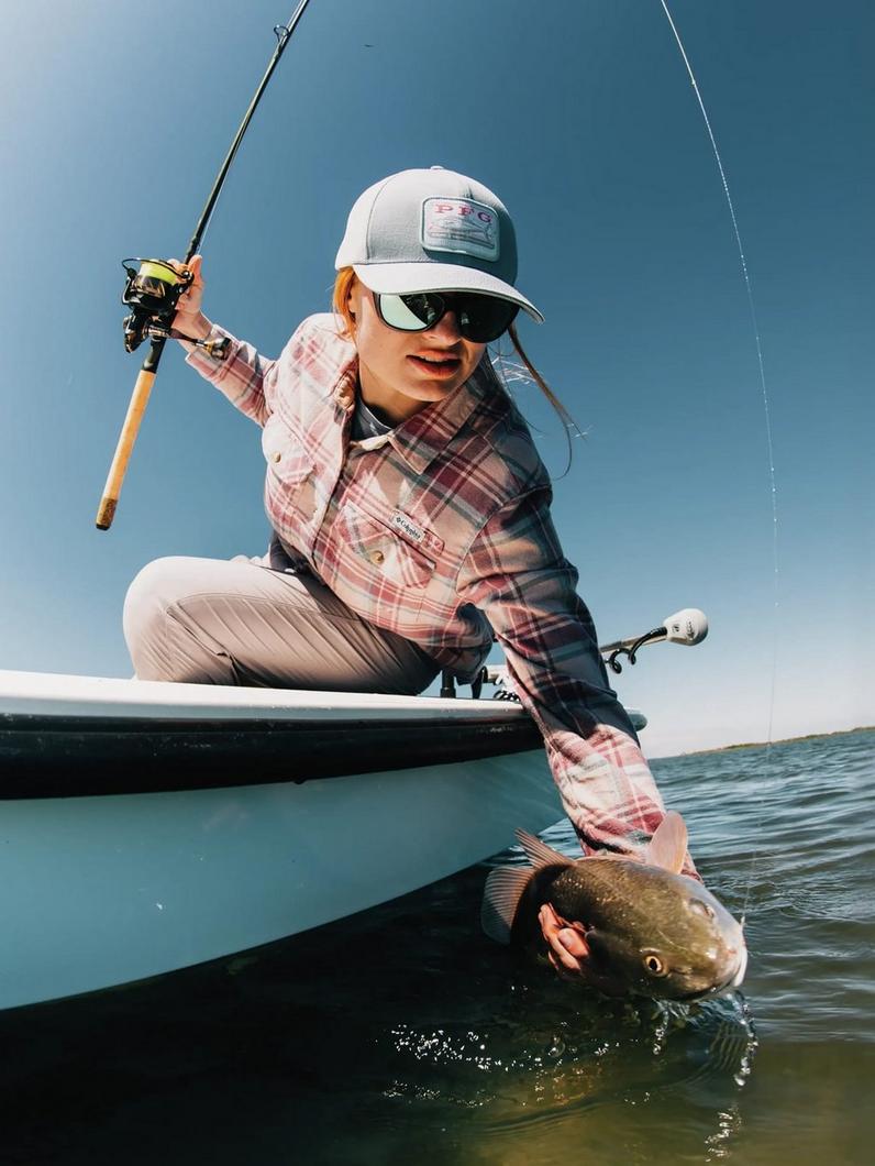 Woman in plaid fishing shirt and white cap releasing a fish from boat while holding fishing rod.