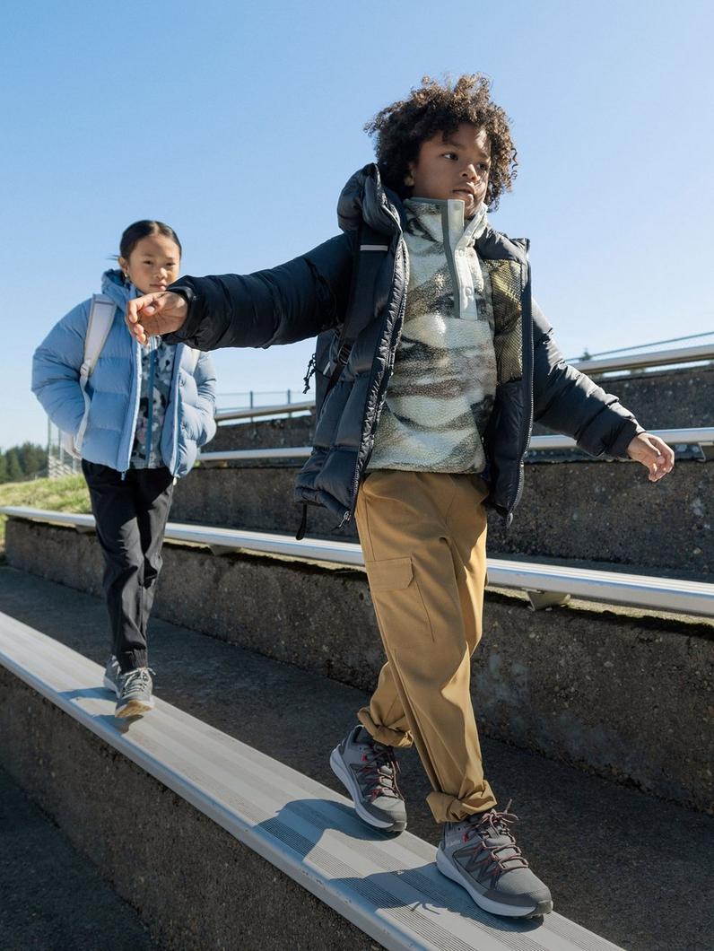 Child balancing on outdoor beam wearing navy puffer jacket, tan pants, and hiking boots with backpack.