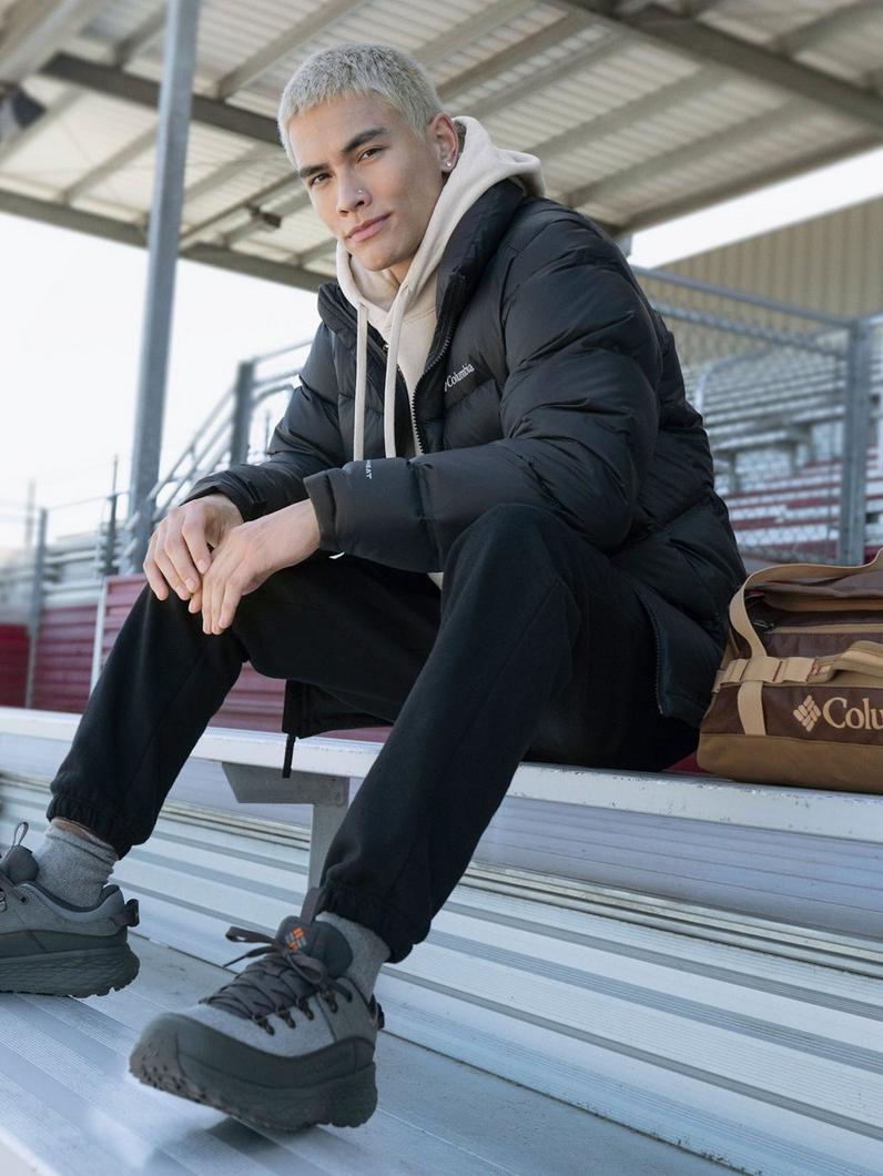 Man sitting on bleachers wearing black puffer jacket layered over beige hoodie with black pants and sneakers.