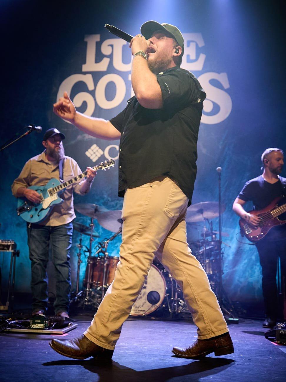 Male performer singing into microphone on stage wearing khaki pants, dark shirt, and cap with band members.