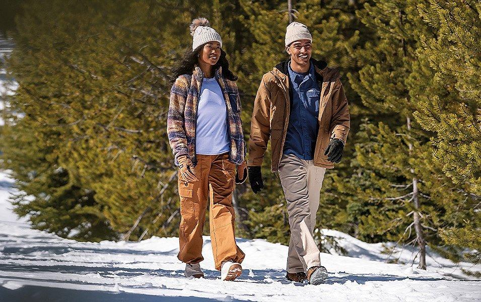 man an woman wearing Columbia Hiking pants walk down a snowy covered path. 