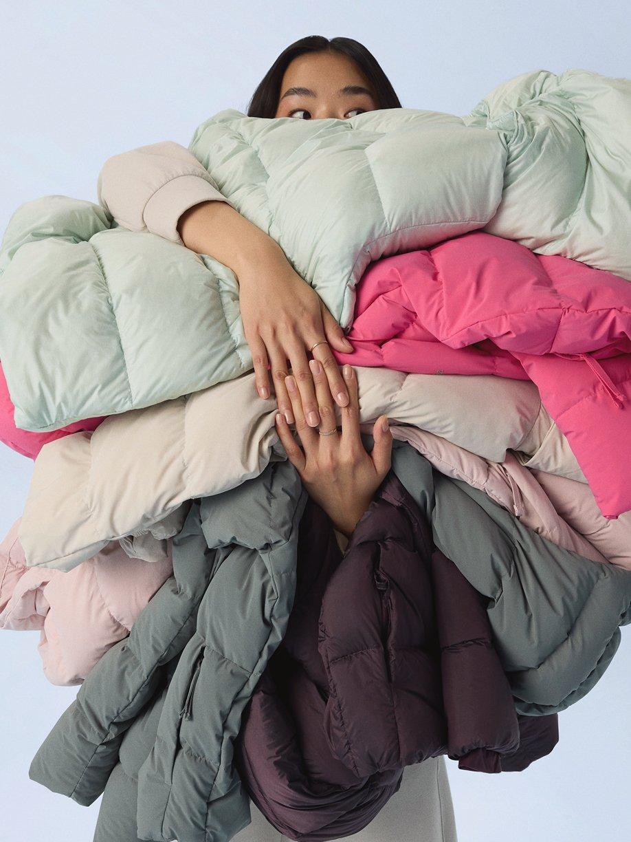 Woman holding an armful of colorful puffer jackets in mint green, pink, beige, and gray tones.
