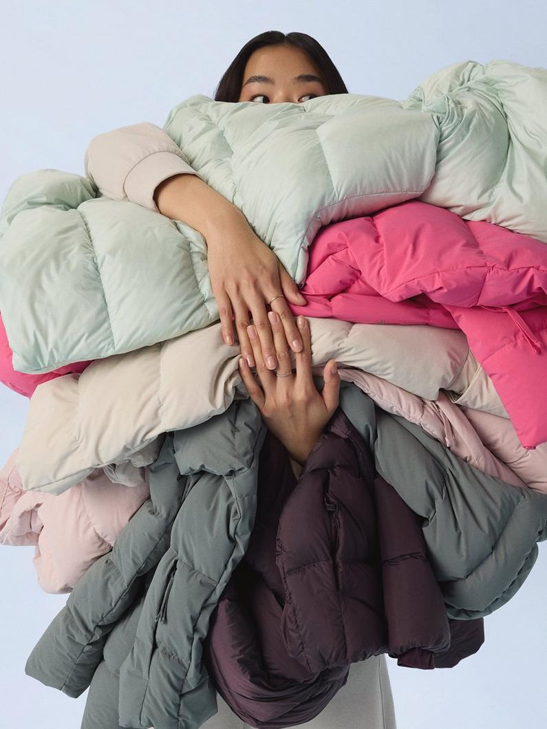 Woman holding an armful of colorful puffer jackets in mint green, pink, beige, and gray tones.