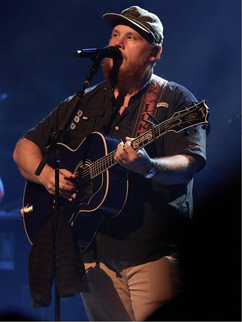 Bearded man wearing a cap singing into a microphone while playing an acoustic guitar on stage.