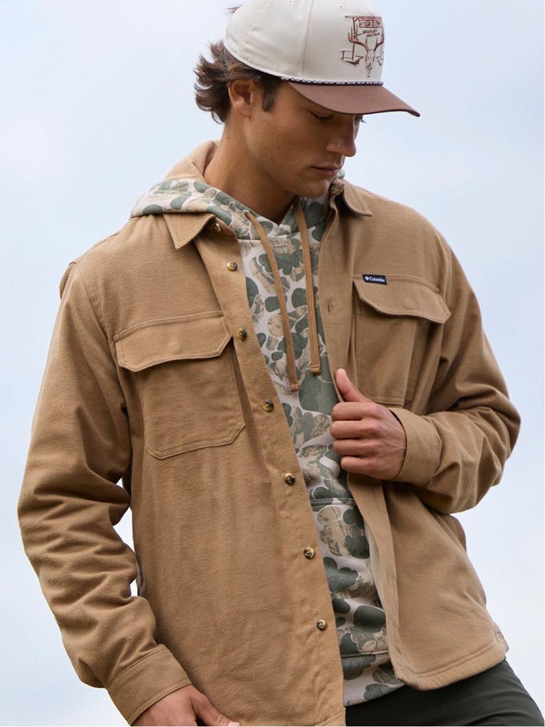 Man wearing tan fleece shirt jacket over camouflage hoodie and baseball cap, looking down.
