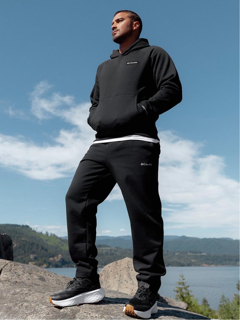 Man standing on rocky outcrop wearing black hoodie and joggers with athletic shoes overlooking a lake.