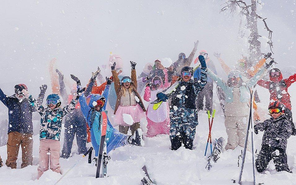 More than a dozen skiers, many in costumes, stand by their skis on a snowy day, with arms raised in celebration.