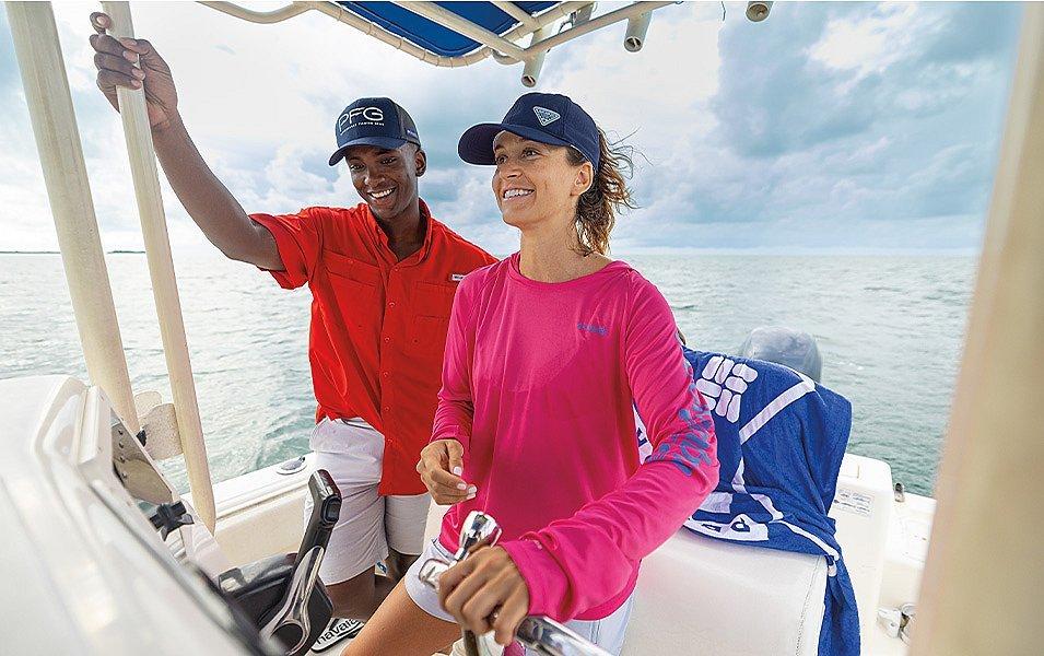Two smiling individuals stand on the deck of a boat; the woman is at the helm. 