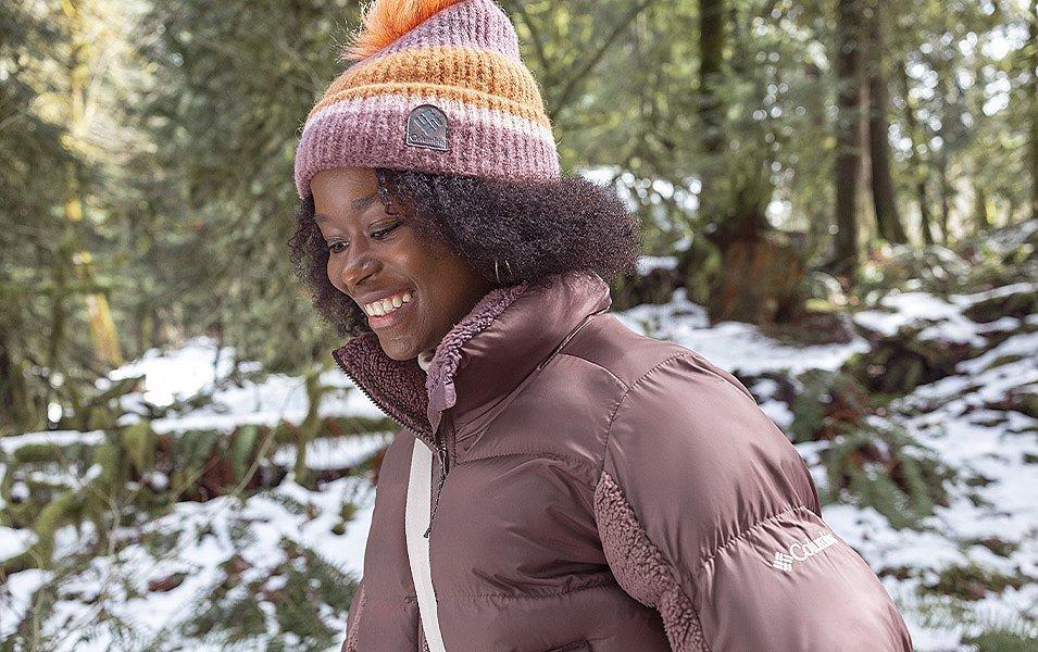 A smiling woman wearing a puffy jacket and knit hat is walking through a snow covered forest.