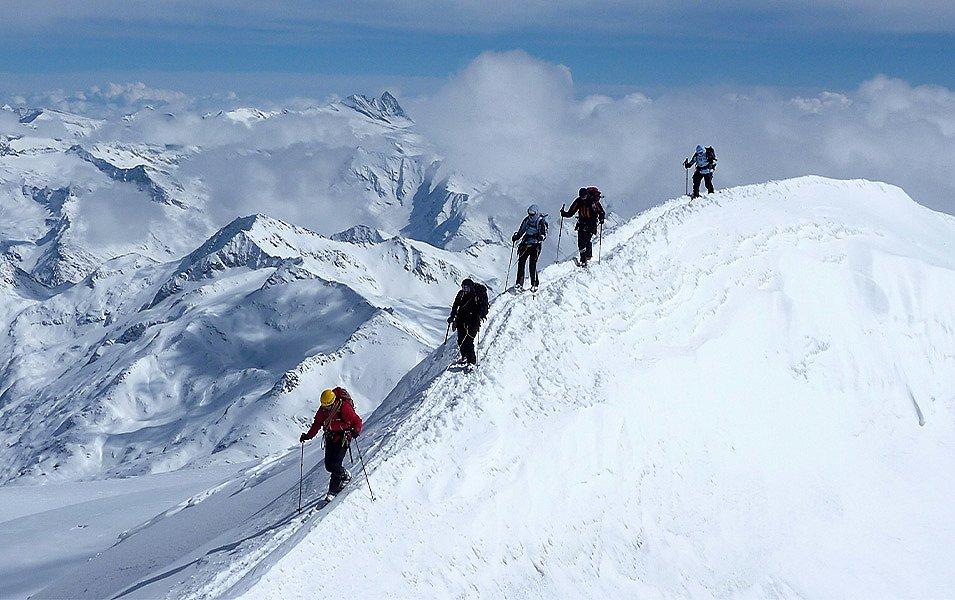 Five people wearing snowshoes and using ski poles for balance make their way down a steep, snowcovered ridge.