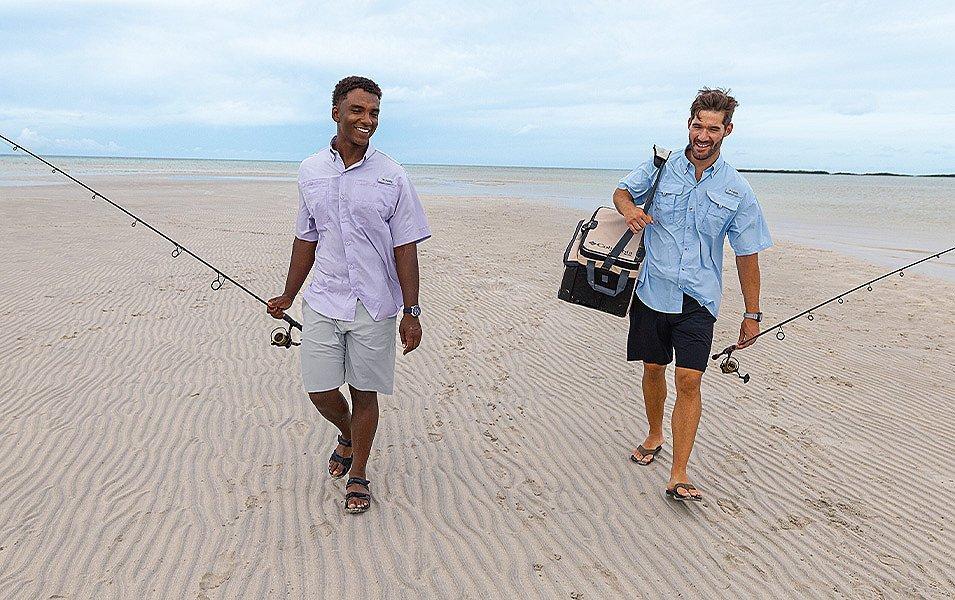 Two men in shorts and sandals are carrying fishing poles as they walk along a sandy beach.