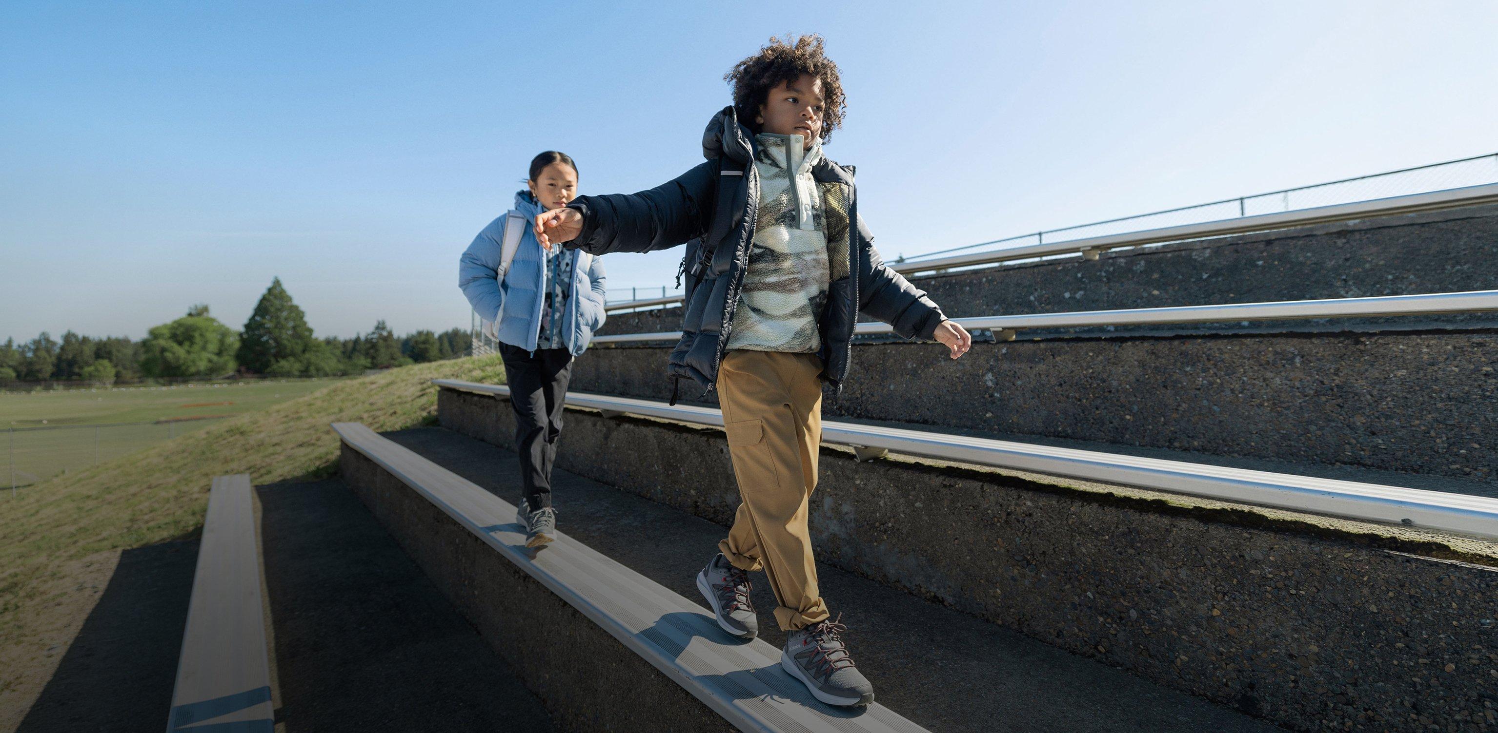 Two children walking down outdoor steps wearing puffer jackets, hoodies, and sneakers with backpacks.