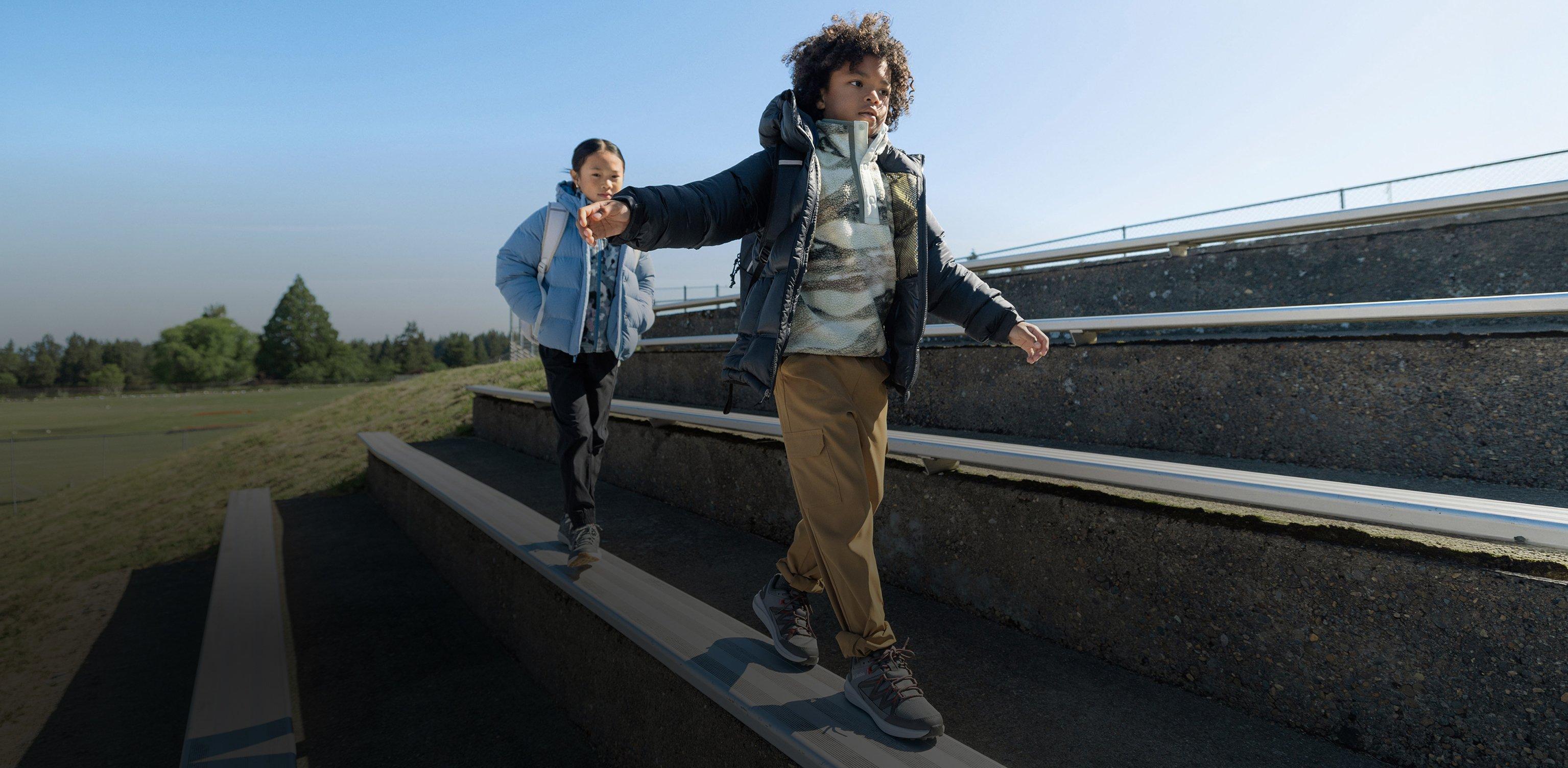 Two children balancing on outdoor concrete steps wearing puffer jackets and sneakers.