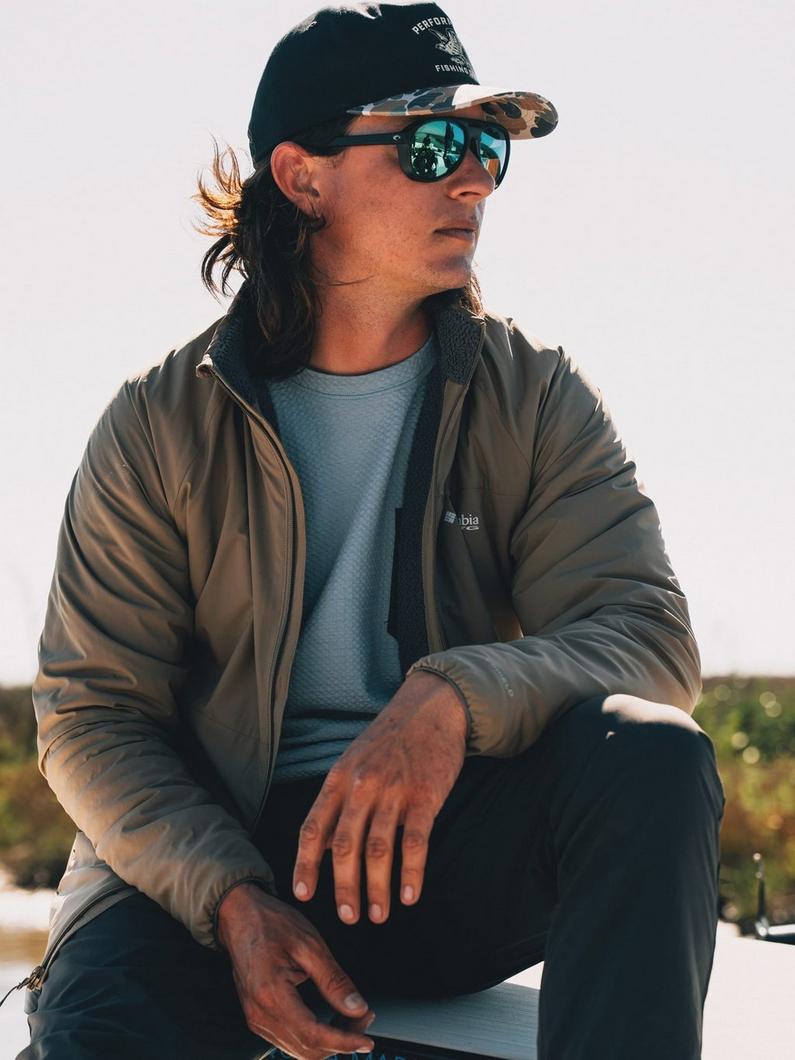 Person wearing mirrored sunglasses, black cap, olive jacket over blue shirt, sitting outdoors.