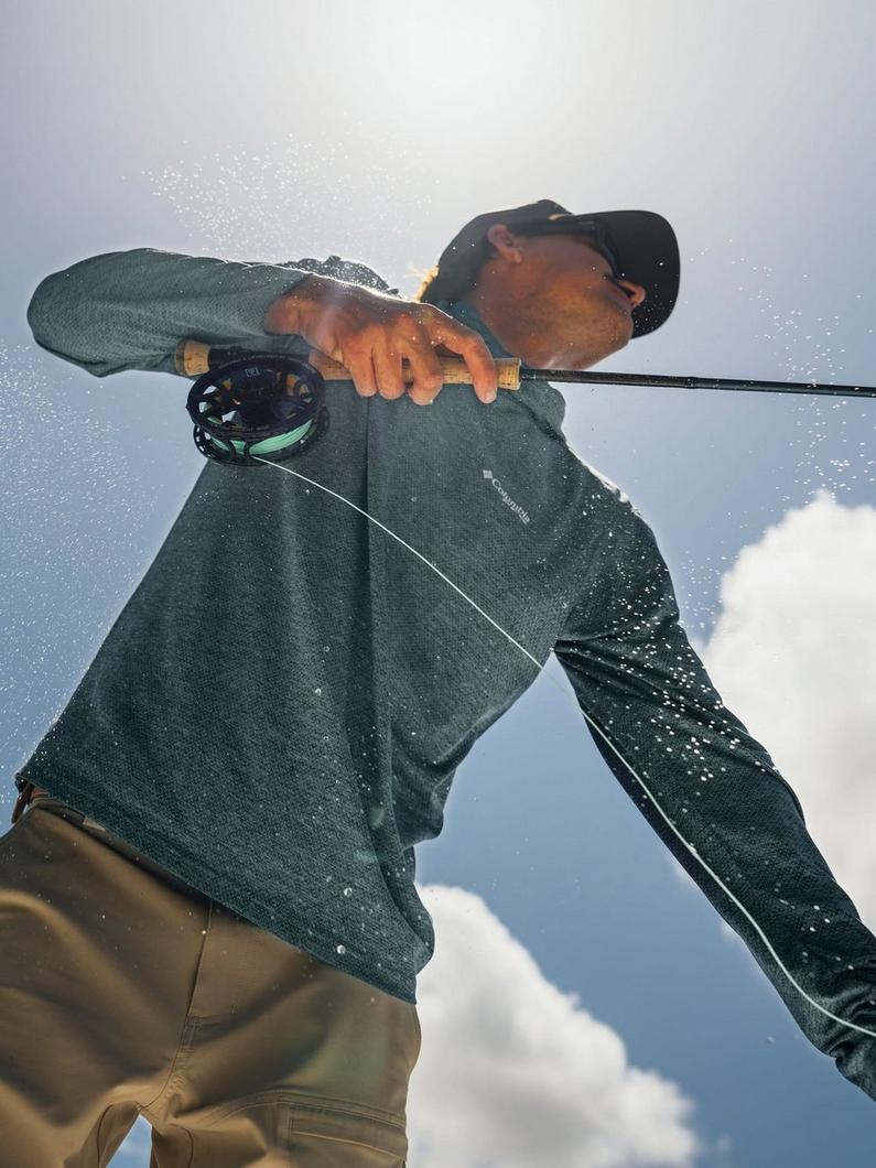 Man in dark green jacket and cap casting a fly fishing rod with water droplets spraying around him.