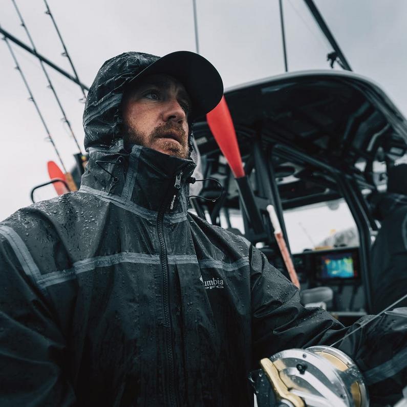 Man wearing waterproof jacket and cap while fishing on a boat in wet weather conditions.