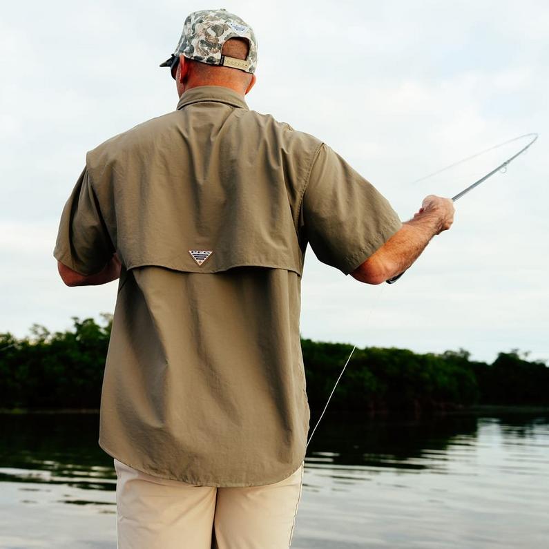 Man wearing tan fishing shirt and camouflage cap casting a fishing rod by the water.