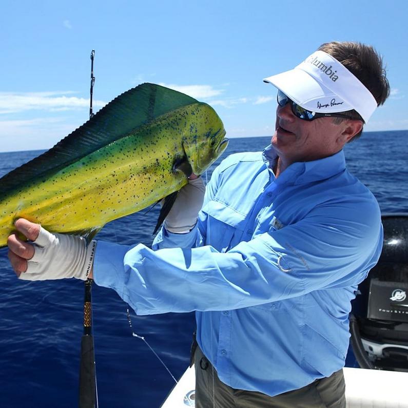 Man wearing white visor and blue fishing shirt holding a large yellow and green mahi-mahi fish on a boat.