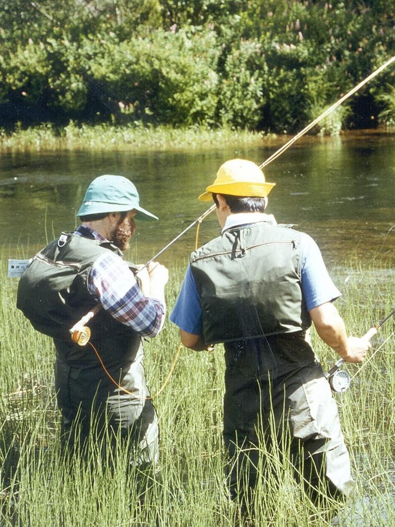 Two people fly fishing in waders and sun hats along a riverbank surrounded by grassy vegetation and trees.