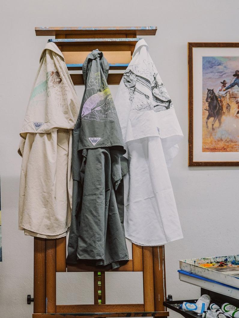 Three shirts from the PFG Artist Series hanging on a hook.