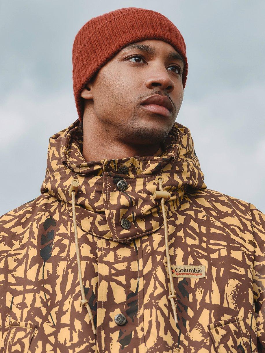 Man wearing rust-colored ribbed knit beanie and camouflage patterned jacket with hood.