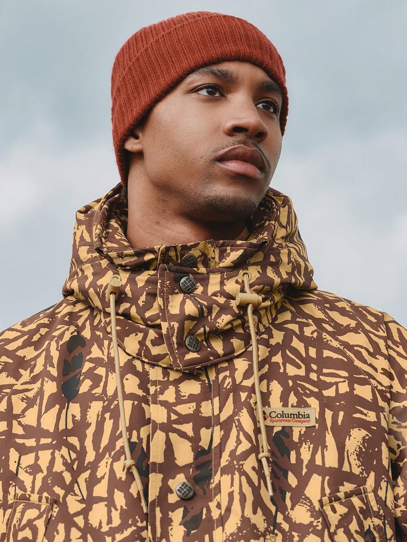 Man wearing rust-colored ribbed knit beanie and camouflage patterned jacket with hood.