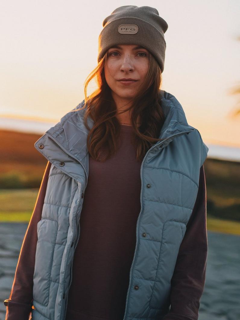 Woman wearing gray knit beanie and blue quilted vest outdoors at sunset.