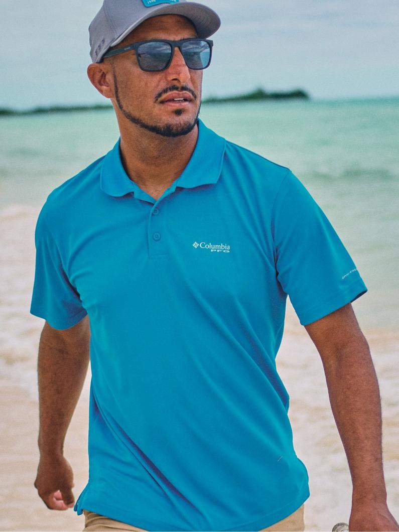 Man wearing turquoise polo shirt, gray cap, and sunglasses standing on a beach.