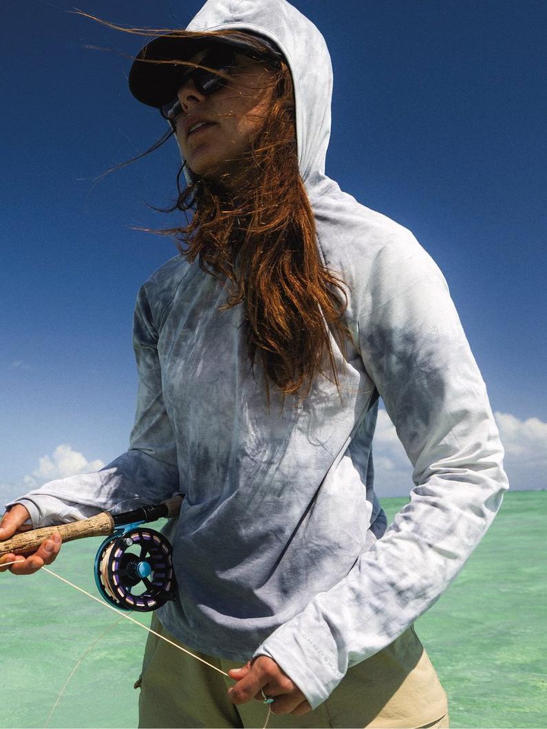 Woman fly fishing in shallow turquoise water wearing white hooded sun protection shirt and black cap.