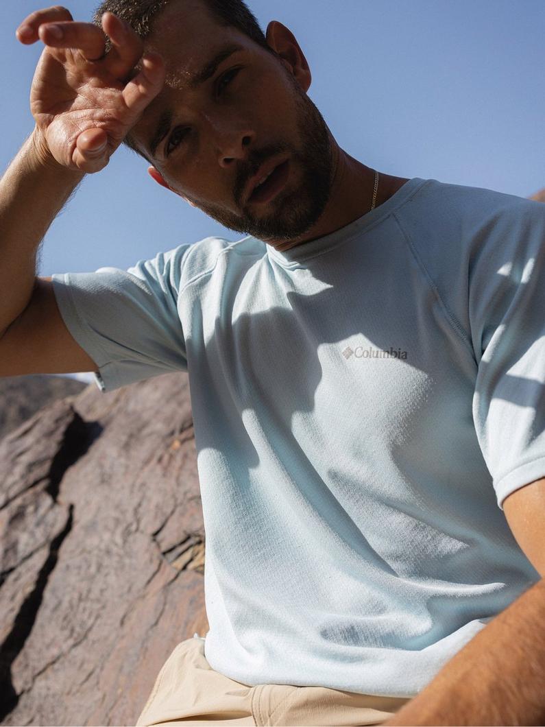 Man shielding eyes from sun while wearing light blue performance shirt outdoors on rocky terrain.