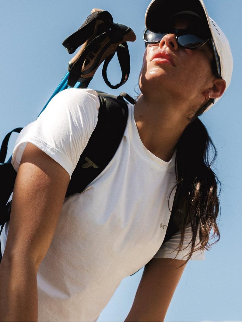 Woman in white t-shirt and cap holding golf glove, wearing sunglasses and carrying golf club bag.