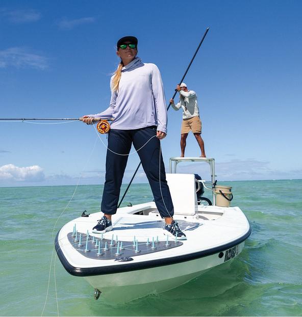 Woman in white long-sleeve shirt and navy pants standing on small fishing boat holding rod with man poling behind.
